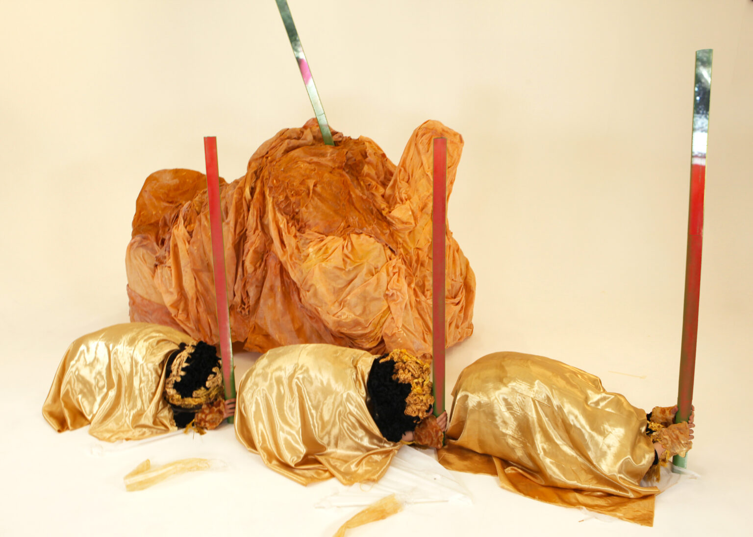 Three performers with golden yellow satin fabrics covering their bodies are crouching on the floor while holding the tubes to stand. The tube has the projected imagery of red liquid moving up and down. The winkled tawny sculpture with the tube penetrating from its back is placed behind the performers.