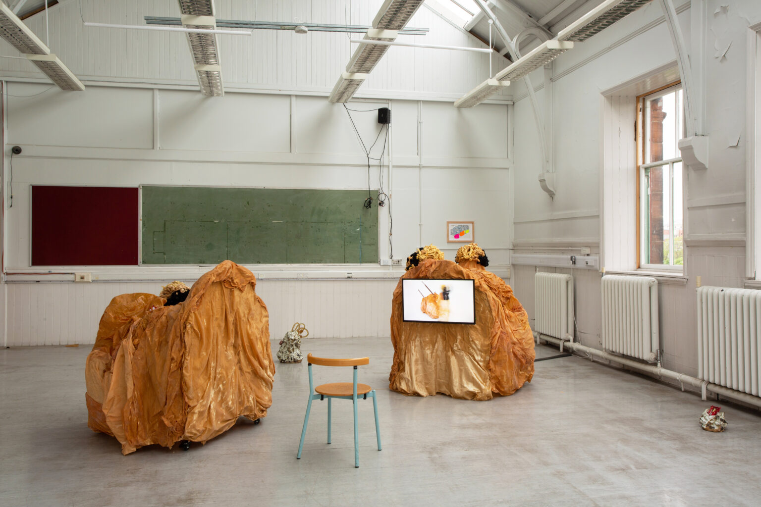 Two big wrinkled tawny-coloured sculptures in the picture have black and yellow masks on the top. One of the sculptures has a monitor installed. In that monitor, the same sculpture but in the form of the two pieces combined is in the middle of the space and a performer is moving around the sculpture with the yellow and black mask on. There are two ceramics on the floor, one of which has yellow transparent flowers and another one has snacks inside. One drawing is installed on the wall.