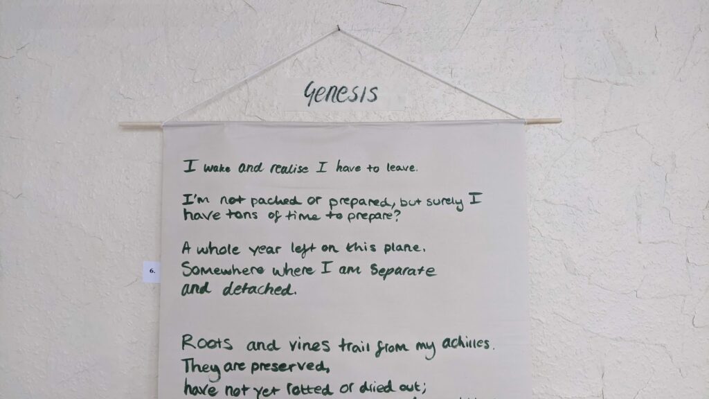 An image of a hanging hand-written text piece. The piece is hung by a dowel, which is strung up with yarn onto a nail in the wall. The wall is a bumpy, wood-chip texture that is painted magnolia. The piece is written in olive-green ink. A large piece of masking tape is above the piece, and reads: "Genesis"