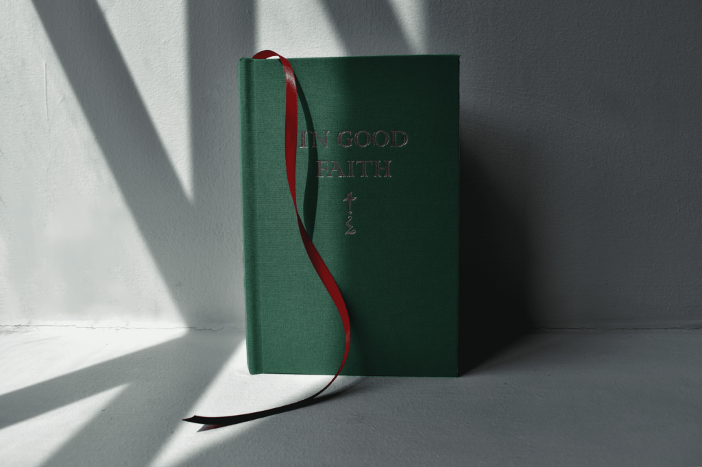 Green book with gold lettering that reads 'In Good Faith' with a glyph underneath and a red ribbon. The book is photographed in natural light and there are diagonal shadows from a nearby window.